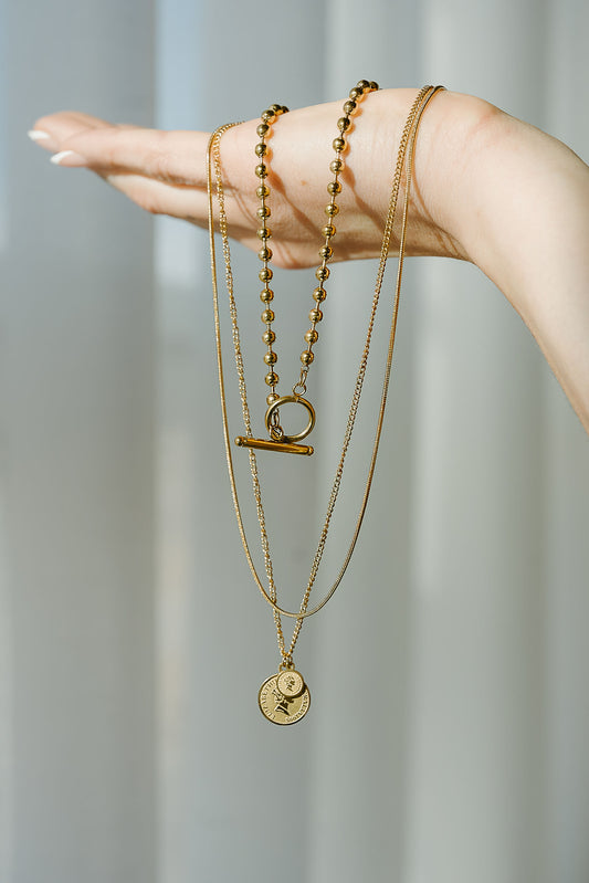 Hand wearing multiple gold necklaces with various pendants against a neutral background