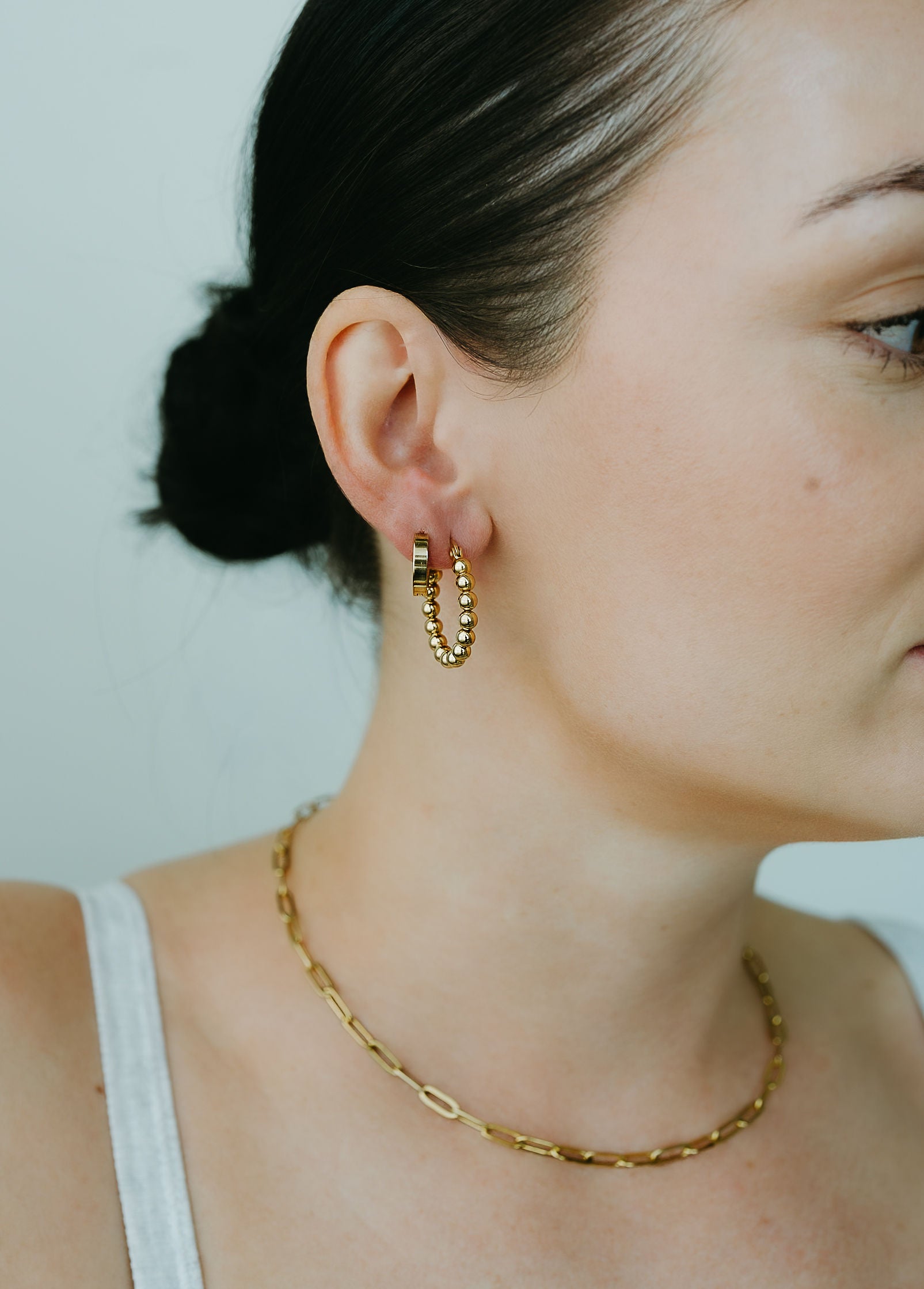 Close-up of a person wearing gold hoop earrings and a gold chain necklace against a neutral background.