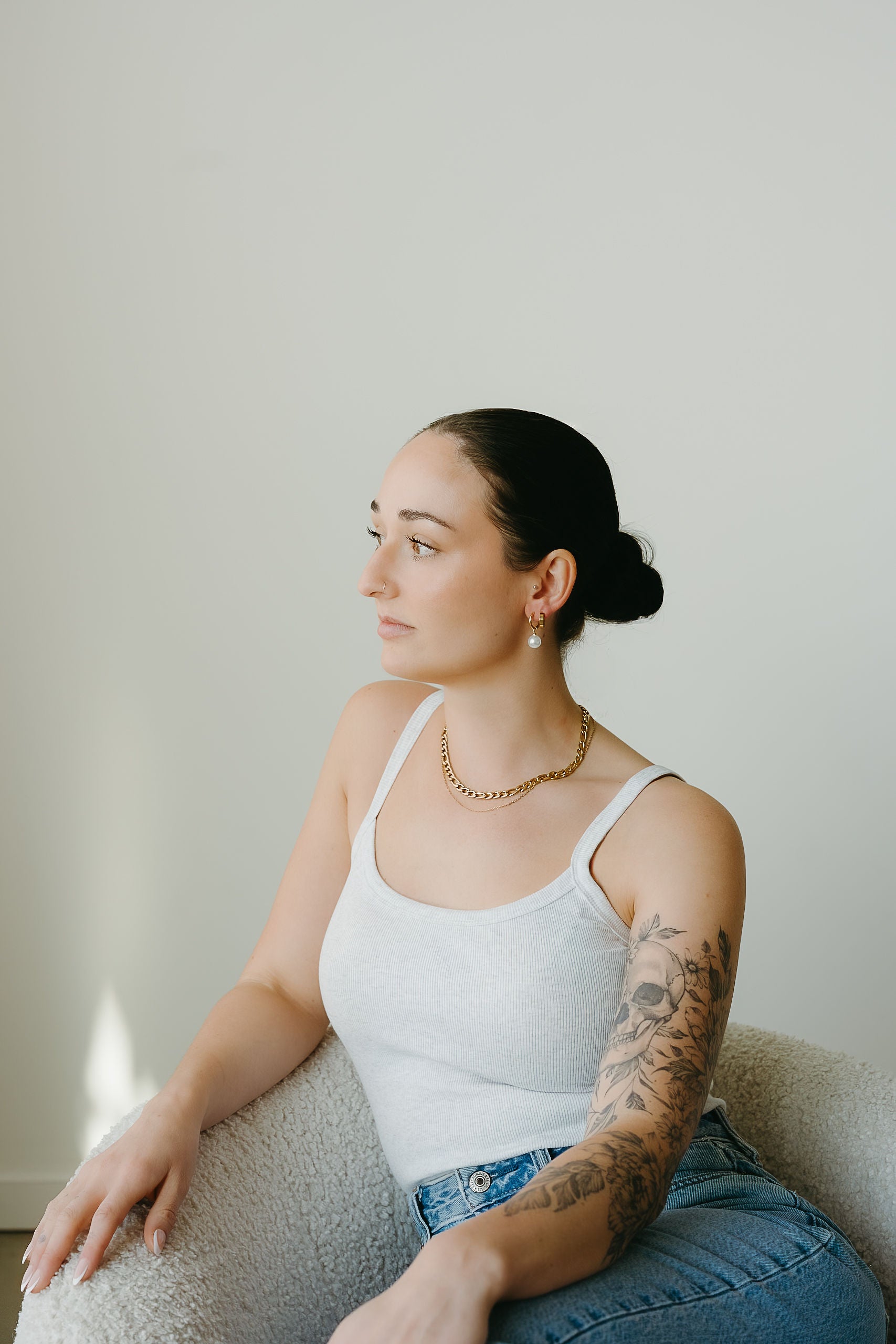 Woman sitting on a couch wearing a white tank top and blue jeans, with a neutral background.