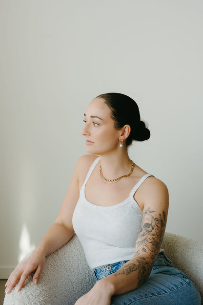 Woman sitting on a couch wearing a white tank top and blue jeans, with a neutral background.