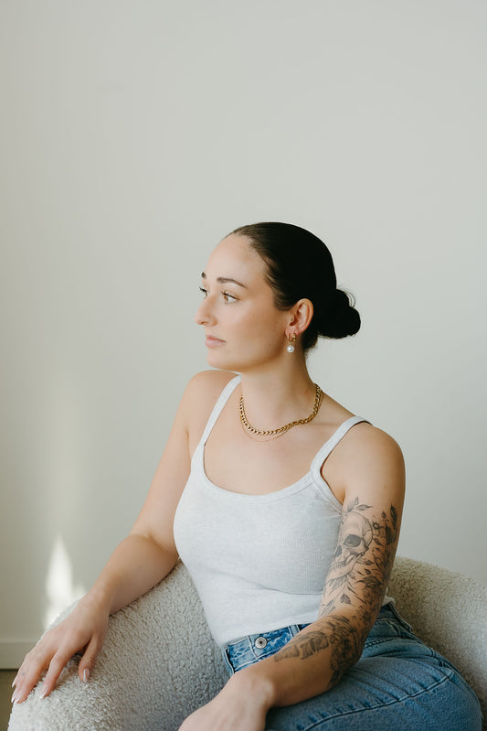 Woman sitting on a couch wearing a white tank top and blue jeans, with a neutral background.
