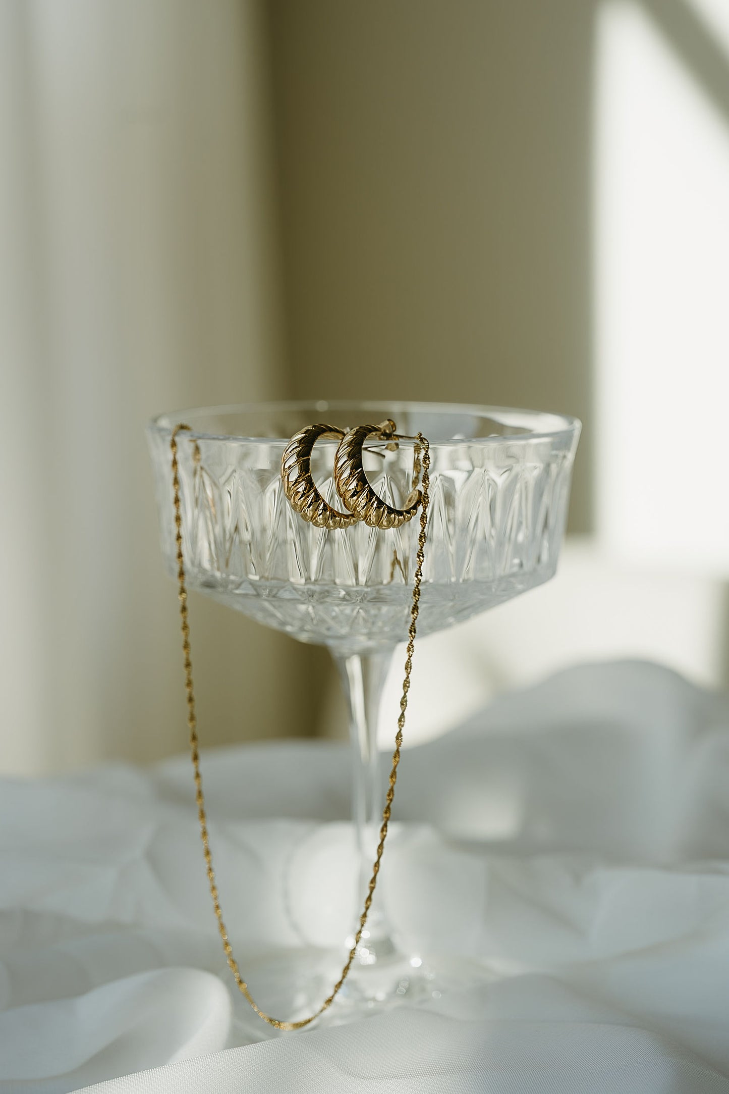 Gold hoop earrings hanging from a gold chain on a clear glass with a white background
